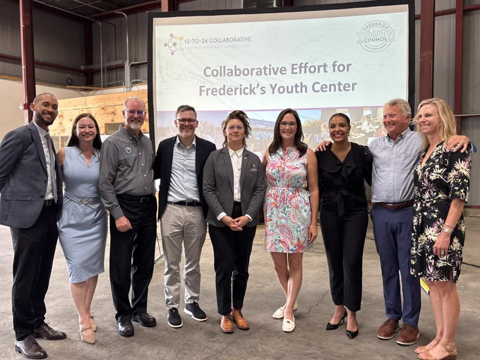 A group of nine people stands together in an industrial warehouse space with concrete flooring, tall metal walls, and exposed beams. They are positioned in front of a large projection screen displaying a presentation slide that reads, “12‑to‑24 Collaborative” with a circular logo, and “Collaborative Effort for Frederick’s Youth Center,” along with the Frederick Youth Council logo in the upper right corner. The group is dressed in a mix of business and semi‑formal attire, including patterned dresses, tailored shirts, jackets, slacks, and dress shoes. They stand side by side in a loosely posed line. Behind them, portions of a wooden structure, industrial equipment, and warehouse columns are visible.