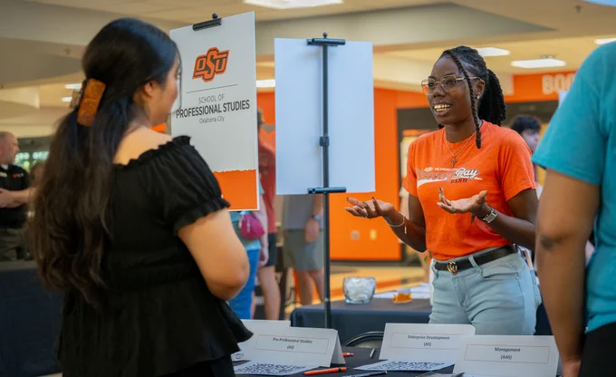 An OSU-OKC staff member speaking with an enrollee. She wears an orange shirt, glasses, and stands behind a booth beside a sign that reads "School of Professional Studies."