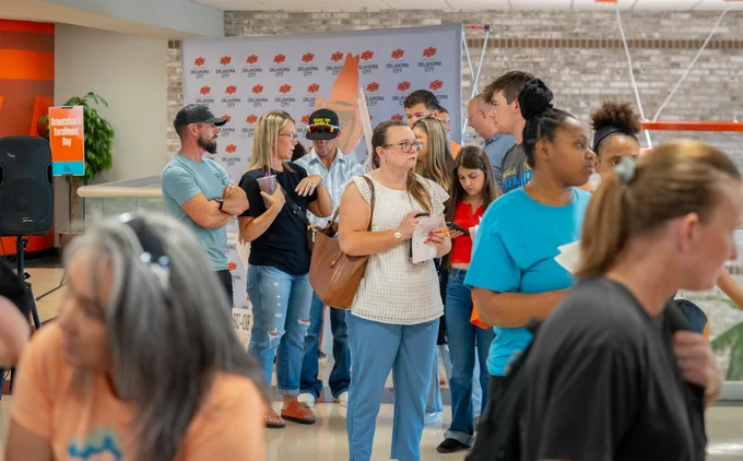 A line of campus visitors and enrollees waiting to speak to the Academic Advising booth. They all stand in front of a photo backdrop, grey with OSU logos printed on.