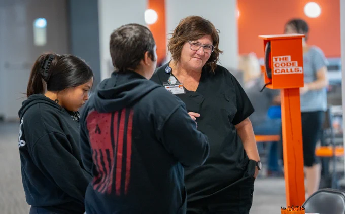 Two people speaking with a woman part of the Health Science school. They all wear black, but the staff member wears glasses and her name tag is clipped to her collar. The "Code Calls" phone is just behind them.