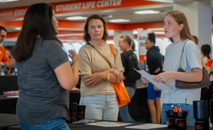 A mother and a daughter speaking with a woman in Career Services. The woman stand behind a booth draped in black table cloth, her back is turned. Behind the visitors is the HUB and other students receiving information from other tables.