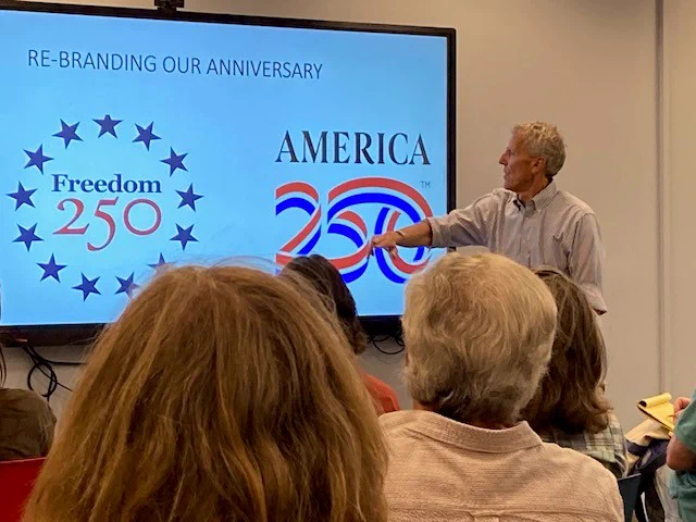 Historian Karl Brooks stands in front of an audience looking at a screen that reads "Re-branding our anniversary" with the logos for Freedom 250 and America 250.
