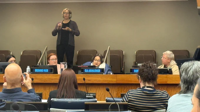 A formal meeting room with a panel seated behind a long desk, each with microphones and nameplates. A speaker at the center reads from notes while others listen. In front, attendees sit in rows, some using devices. Behind the panel, a raised platform holds a sign language interpreter actively signing. The setting is structured and well-lit, with conference seating and interpretation to support accessibility.