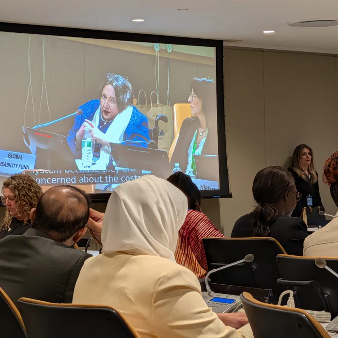 A conference room seen from the audience. People sit facing a large screen showing a close-up of a speaker at a panel, with captions visible. The speaker sits at a desk with a microphone and nameplate. On the right side of the room, a sign language interpreter stands beside the screen. Tables have built-in microphones and control panels, and the lighting is bright and even.