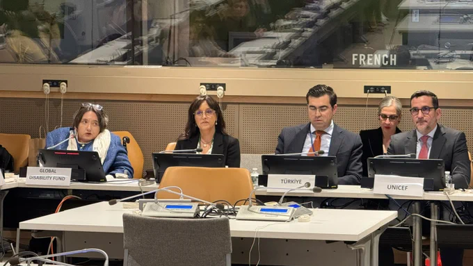 A formal conference setting with participants seated at a long table equipped with microphones and digital panels. Nameplates read “Global Disability Fund,” “Türkiye,” and “UNICEF.” Multiple speakers face the room while attendees sit nearby. Behind them, a glass booth labeled “French” indicates interpretation services. The room is well-lit and arranged for multilingual, structured discussion with technical equipment and cables visible on the table.