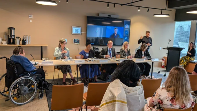 A panel discussion in a modern meeting room. Seven speakers sit behind a long table with microphones and nameplates, while attendees listen from rows of chairs. One of them uses a wheelchair at the left. A large screen behind shows a remote speaker. On the right, a sign language interpreter stands near a podium. Coffee service sits on a side counter, and ceiling lights illuminate the space evenly.
