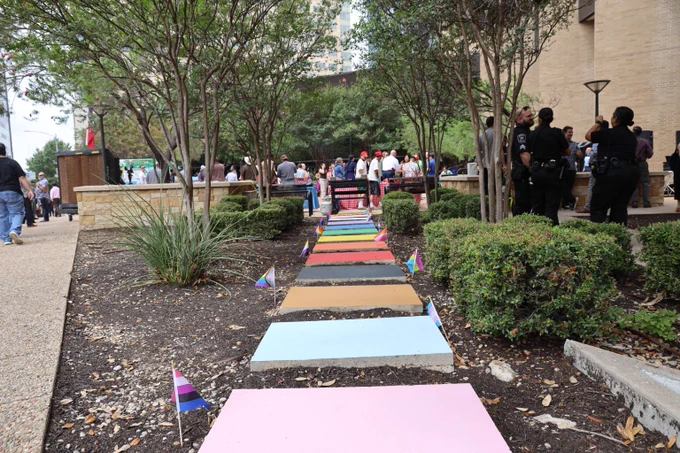 Colorful painted concrete slabs form a rainbow pathway lined with small pride flags in an outdoor urban garden area.
