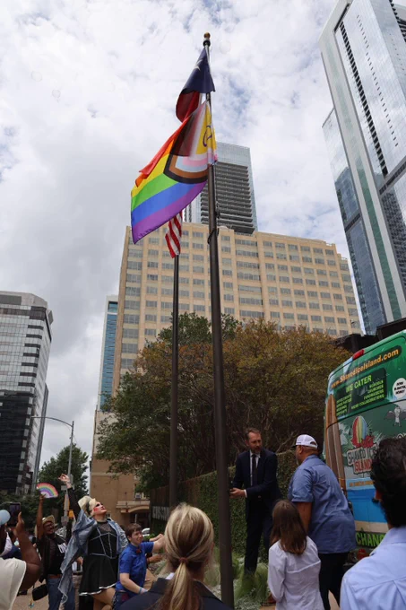 Rainbow pride flag and American flag flying on poles at 700 Lavaca Street in Austin, Texas, with people gathered around.