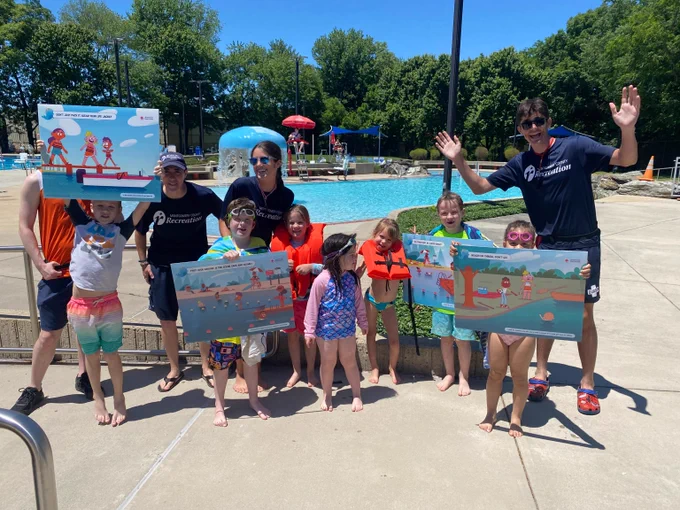 Group of children and adults holding swimming safety posters by an outdoor pool on a sunny day.