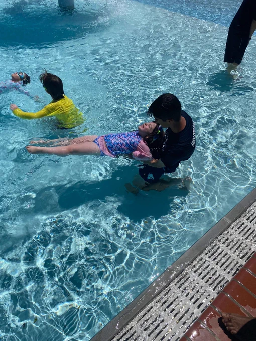 A child in a pink and blue swimsuit floats on their back in a pool, supported by an adult in a black swim shirt.