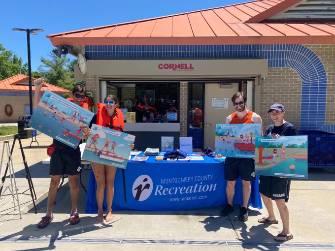 Four people stand in front of a Montgomery County Recreation booth holding colorful swimming safety posters on a sunny day.