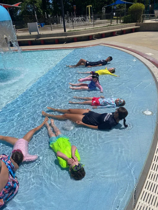 Children lying and floating in shallow pool water on a sunny day at outdoor pool.