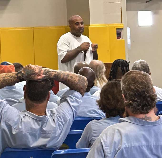 A man in a white shirt speaking to a seated group of people wearing light blue uniforms in a room with yellow padded walls.