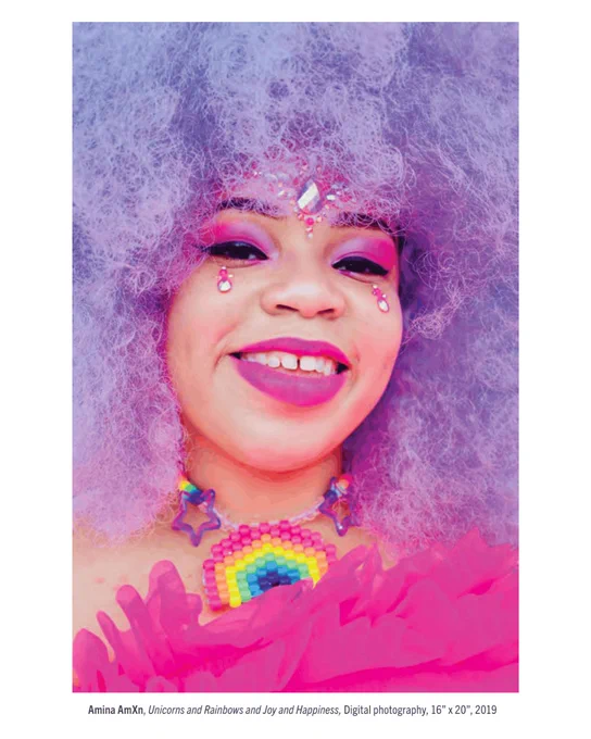 Portrait photograph of a smiling person with voluminous lavender-gray curly hair, pink and purple makeup, jeweled face decorations, and a rainbow beaded necklace. Bright pink fabric fills the foreground, creating a vibrant, celebratory look.