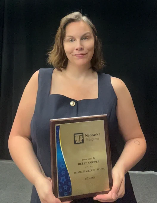 East teacher Helen Cooper stands in the school auditorium with her plaque for winning the 2025-26 Nebraska Thespians Theater Teacher of the Year Award. She is the school's theater director. She is holding the plaque in front of her with both hands.