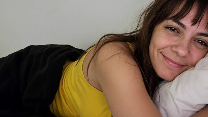 Porträt einer lächelnden Frau mit dunklen Haaren und Pony. Sie liegt seitlich im Bett, trägt ein sonnengelbes Tanktop und hat den Kopf auf ein weißes Kissen gebettet. Eine schwarze Kuscheldecke liegt über ihr.