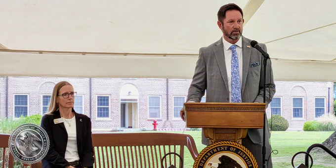 A person in a suit stands at a podium with the "Bureau of Prisons" seal, speaking into a microphone under a tent. Another individual is seated nearby. The backdrop includes a brick building and lawn area.