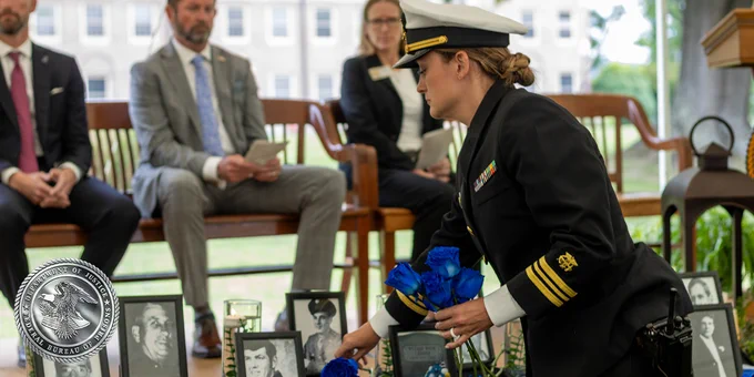 A uniformed individual places blue roses at a memorial with black-and-white photographs on display. In the background, several people are seated on a bench, observing the ceremony. 