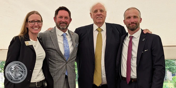 Four individuals are standing together under a tent, dressed in formal attire. They are smiling at the camera. The group includes well-dressed figures with a visible emblem that resembles the Federal Bureau of Prisons.