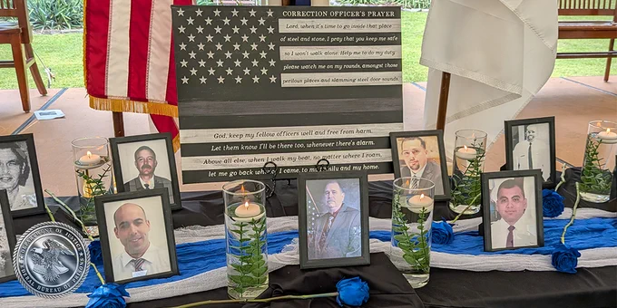 This image shows a memorial display honoring correction officers. In the center is a "Correction Officer's Prayer" sign with a flag design. Surrounding it are framed photos of individuals, each accompanied by a single blue rose and a lit candle in glass holders. The display includes the American flag and other ceremonial elements.
