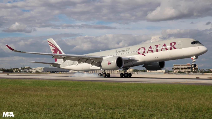 A Qatar Airways airplane is landing on a runway. The aircraft is painted in white with the Qatar Airways logo. The sky is partly cloudy, and there are buildings in the background.