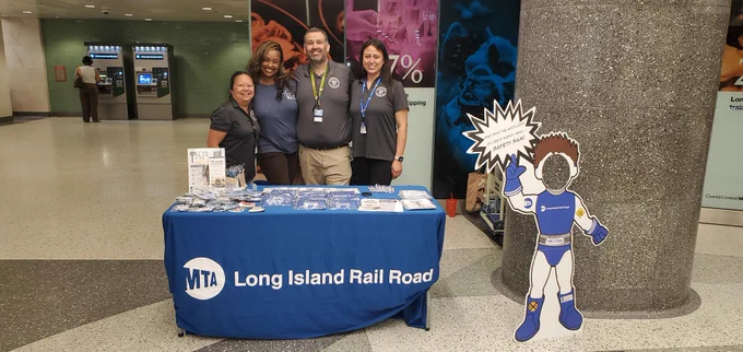 A group of four people standing behind a blue Long Island Rail Road TRACKS table, which is displaying informational materials inside the Grand Central Madison concourse.
