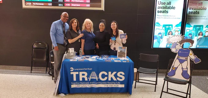 A group of five people standing behind a blue Long Island Rail Road TRACKS table, which is displaying informational materials inside the Grand Central Madison concourse.
