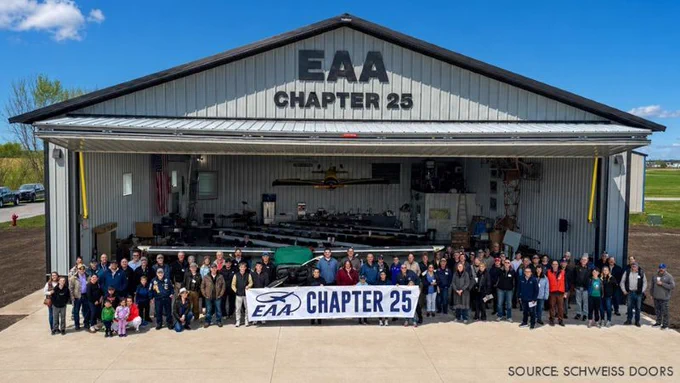 Group of people standing in front of a large metal aircraft hangar with an open bifold door and EAA Chapter 25 signage.