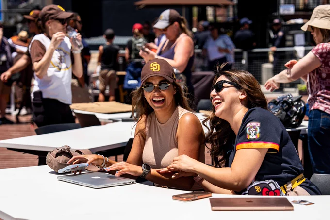 Photo of two Padres fans sitting at a table in Gallagher Square. They are both laughing in the photo. They have their laptops and cell phones with them for Remote Work Wednesdays.