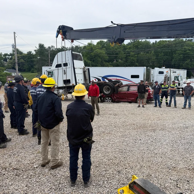 A group of people is gathered around a red car that is overturned next to a large truck with "Godfrey's" logo. A crane is positioned above, and several individuals are wearing helmets and uniforms, suggesting an emergency response or training scenario. There are other trucks and equipment in the background.