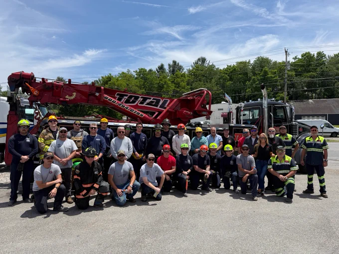 A group of people is gathered outdoors in front of a large red tow truck labeled "Total." They are wearing safety gear, including helmets and reflective clothing. A clear blue sky and trees are in the background.