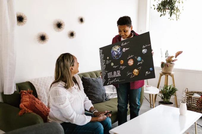 A boy presents a homemade solar system project to his mother in a cozy living room.