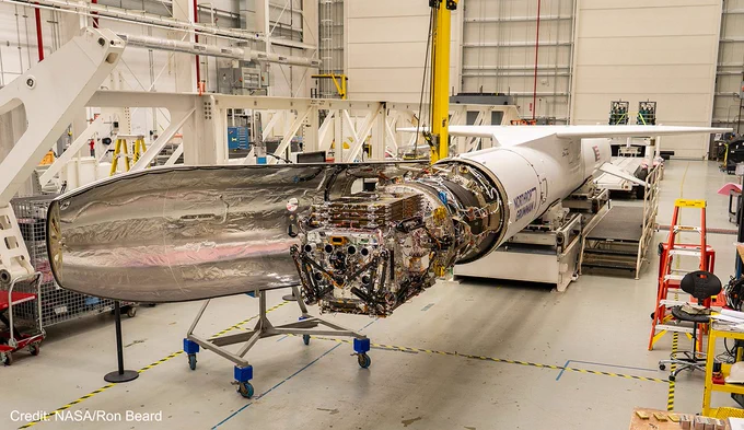 Inside a large white high bay, a small silver satellite is attached to the front of a long white rocket. The satellite has two rows of black lenses on the side facing the camera, two solar panels folded top and bottom, and three robotic arms stowed along the sides and face. The rocket’s nose cone is open, with one side missing and the other folded away like a banana peel, revealing a silvery interior. It has a long wing on the top, and the words “Northrop Grumman” are just visible in blue letters on the right side. This image is watermarked with “Credit: NASA/Ron Beard.”