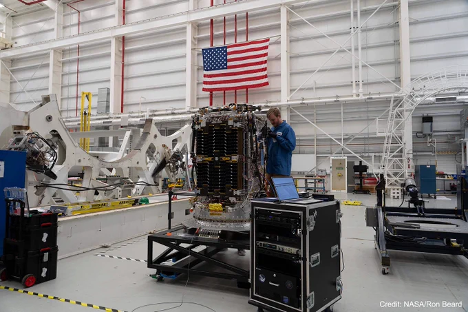 Inside a large white high bay, a man in a blue static discharge jacket stands on a black platform and works on a small satellite. The satellite is silver with black solar panels folded against the near side. A black electronics cart with a laptop on top rests in front of the platform. Other large pieces of equipment are visible to the left and right inside the bay, and an American flag hangs on the far wall. This image is watermarked with “Credit: NASA/Ron Beard.”