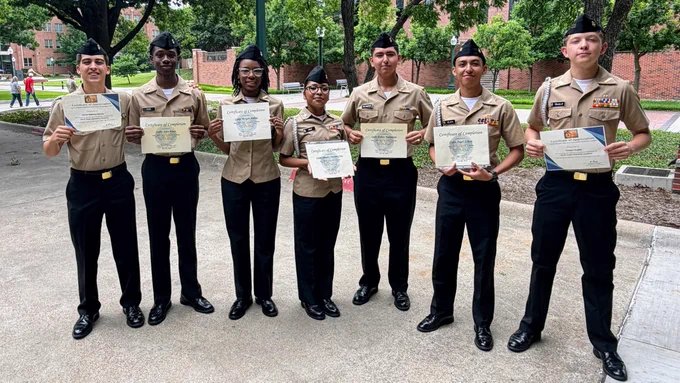 A group of uniformed individuals, likely scouts or cadets, holding certificates or awards in an outdoor setting with trees and buildings.