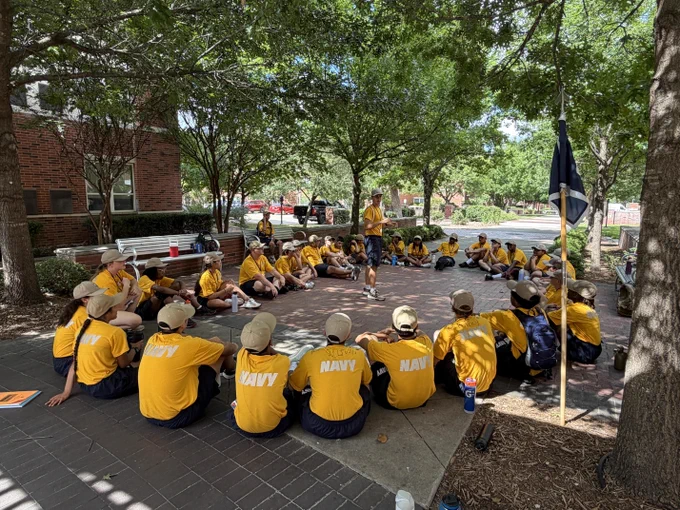 A group of youth in yellow shirts sitting in a circle in a park with trees.