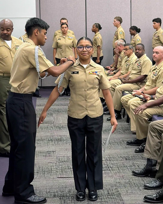 A group of uniformed individuals, some standing and some seated, in a room setting. One individual in the center appears to be wearing glasses and a uniform with patches.