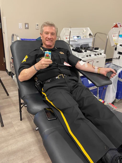 A person in a black uniform is lying on a reclined chair while donating blood. They are holding a small juice carton and smiling. Medical equipment is visible in the background.