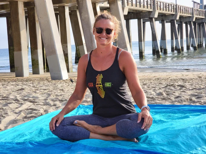 Amy sits cross-legged on a blue mat on the beach, in front of Jennette's Pier. She wearing sunglasses and a sleeveless shirt with a colorful design. The ocean is in the background.