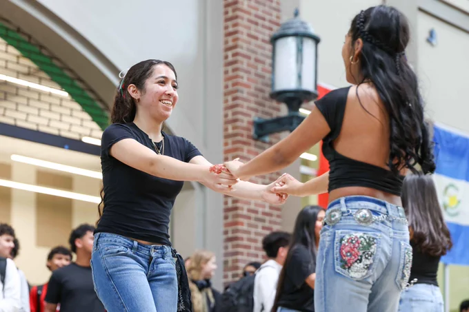 Two women joyfully dance together in an indoor setting. The background features other people and colorful flags.
