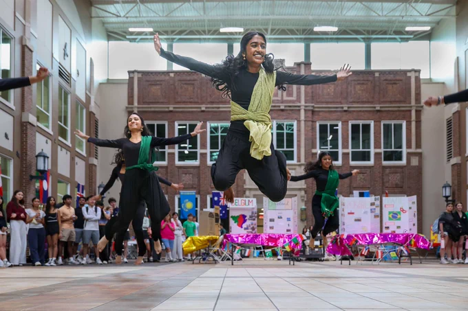 A group of dancers in black outfits and colorful sashes energetically perform a cultural dance in an indoor public space, evoking a sense of joy and unity.