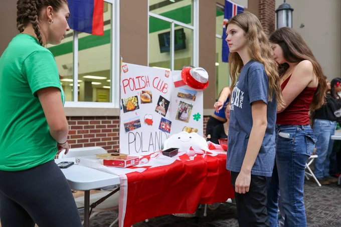 Students stand at a booth for the Polish Club during a school fair, showcasing Polish culture and food. The mood is vibrant and engaging as attendees interact and explore the cultural display.