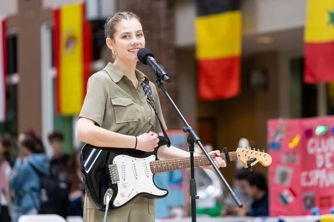 A young woman plays electric guitar and sings into a microphone at a cultural event. Flags and booths in the background suggest diversity and celebration.
