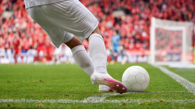 Soccer player in white shorts and socks preparing to kick a white ball near the goal line on a green field.