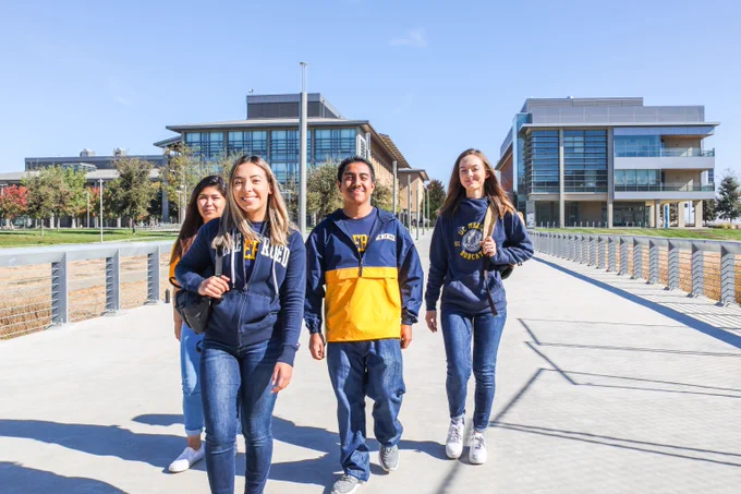 Four students walking on a sunny college campus pathway with modern academic buildings in the background.