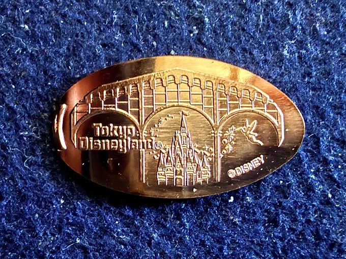 Souvenir medal  ("pressed penny") of the view of the Tokyo Disneyland Cinderella Castle from World Bazaar. Text: "Tokyo Disneyland" ©Disney
