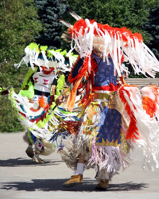 Two individuals are dressed in vibrant, traditional Native American regalia with feathers and colorful patterns, dancing outdoors surrounded by greenery.