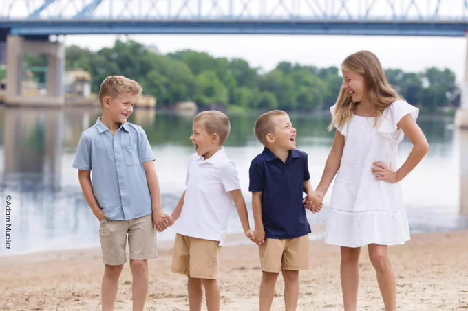A group of four children standing on a sandy beach, holding hands and smiling at each other. There is a bridge in the background over a body of water. Copyright Adam Mueller.
