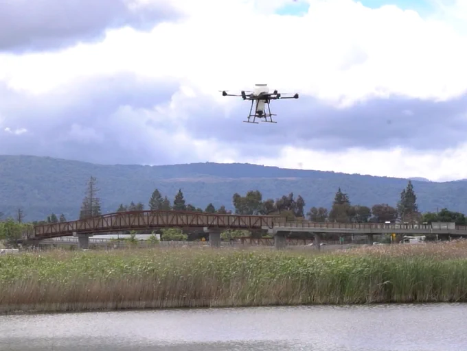a drone hovers over a pond with reeds surrounding it. There is a bridge and foothills in the background.