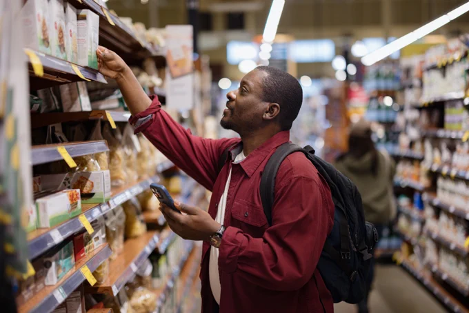 A person in a grocery store aisle reaches for an item on a high shelf while holding a smartphone. They are wearing a red jacket and have a backpack. The shelves are stocked with various packaged goods.